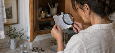 Woman examining an ingredient leaflet with a magnifying glass beside an open medicine cabinet.